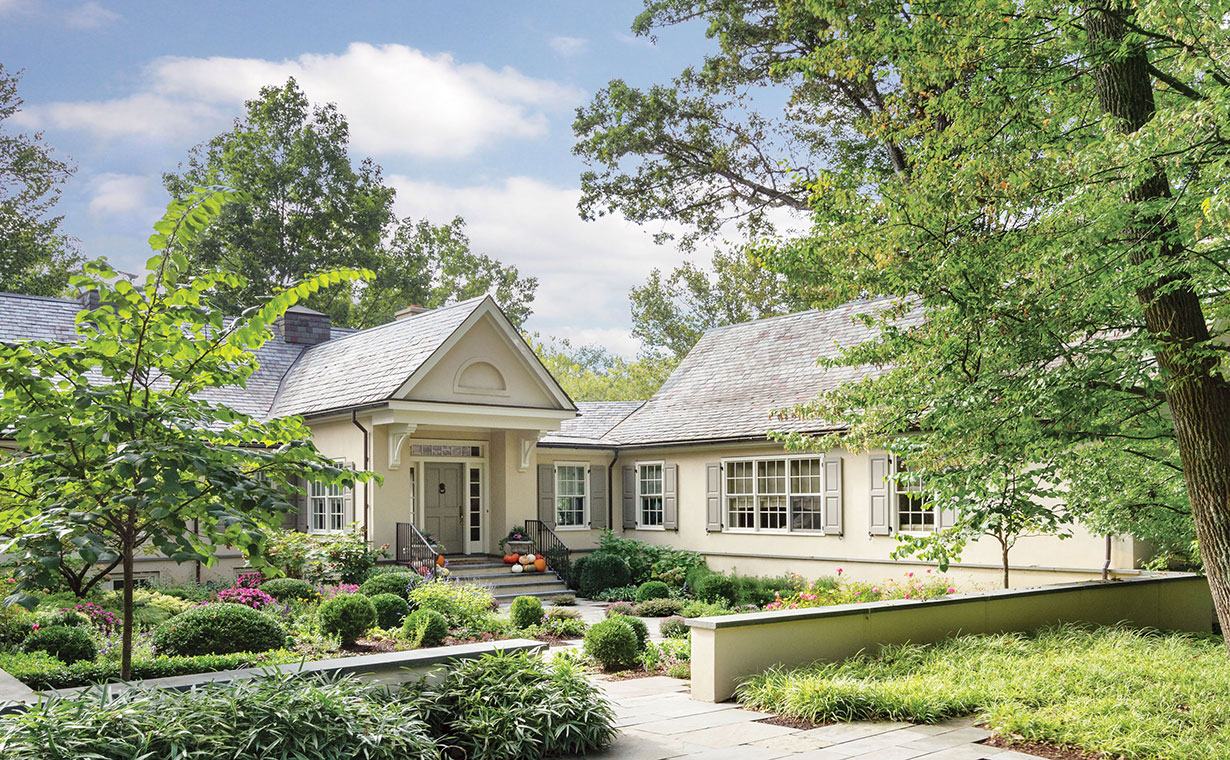 The entry level of this stucco Colonial-style home is highlighted with a bracketed portico and a slate roof that varies in color and proportions. A stucco wall encloses an inner courtyard that in each season is a kaleidoscope of blooming flowers and is punctuated with evergreen for winter.